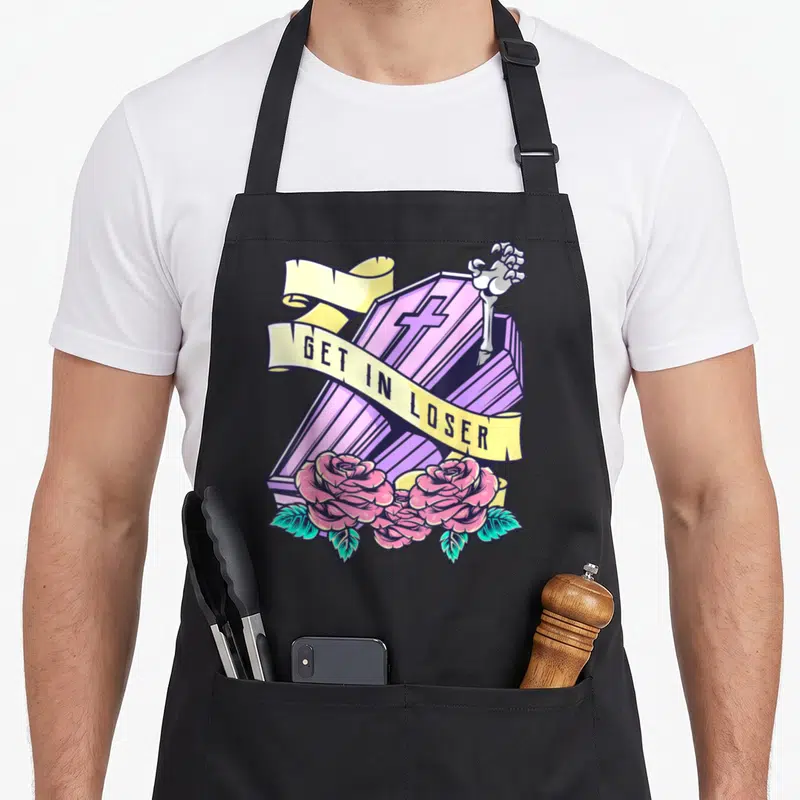Coffin halloween apron - Purple coffin with skeleton hand, yellow banner reading GET IN LOSER, pink roses, teal leaves — shown on a black cotton canvas apron against a white background