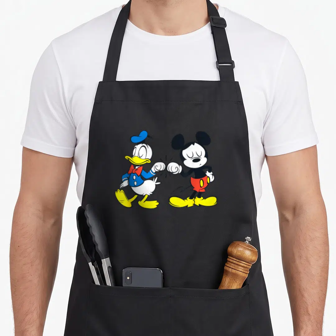 Funny friend duck apron - Donald Duck and Mickey Mouse standing side-by-side performing a fist bump in blue, yellow — shown on a black cotton canvas apron against a white background