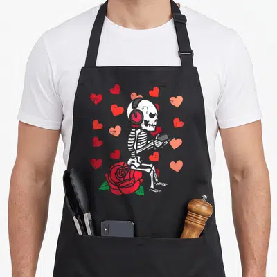 Gamer skeleton valentine's day apron - White skeleton with headphones holding controller sitting on red rose surrounded — shown on a black cotton canvas apron against a white background