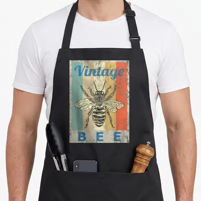 Queen honey bee apron - Retro bee illustration over five vertical stripes with arched Vintage text and B E E below — shown on a black cotton canvas apron against a white background