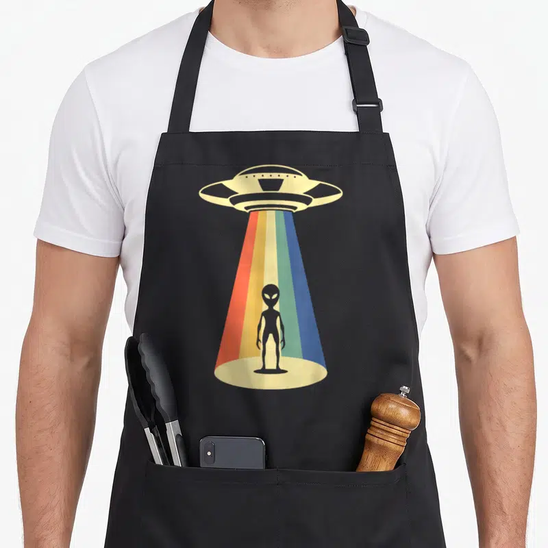 Abduction ufo apron - Grey alien silhouette under UFO rainbow beam on beige base — shown on a black cotton canvas apron against a white background