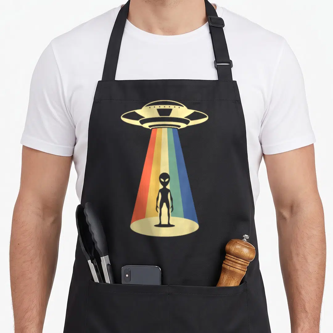 Abduction ufo apron - Grey alien silhouette under UFO rainbow beam on beige base — shown on a black cotton canvas apron against a white background