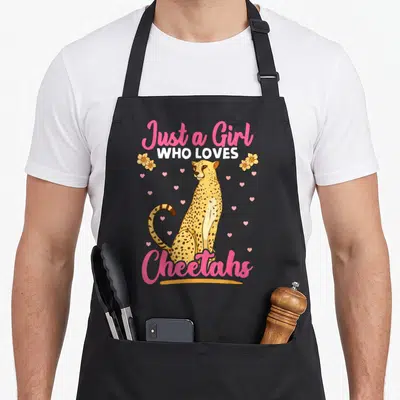 African cheetah apron - Cartoon cheetah sitting between pink Just a Girl Who Loves Cheetahs text with flowers and hearts — shown on a black cotton canvas apron against a white background