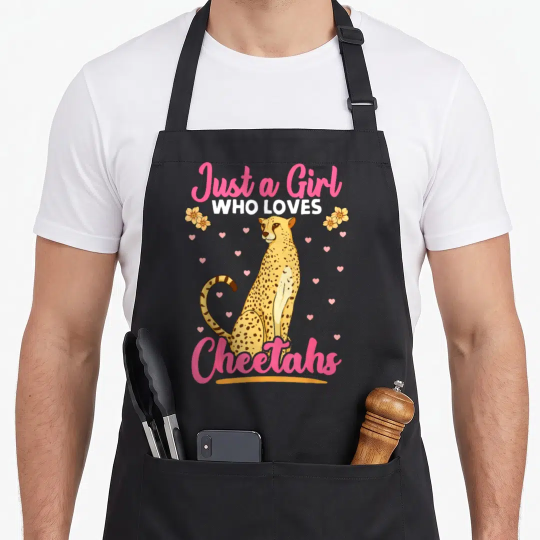 African cheetah apron - Cartoon cheetah sitting between pink Just a Girl Who Loves Cheetahs text with flowers and hearts — shown on a black cotton canvas apron against a white background