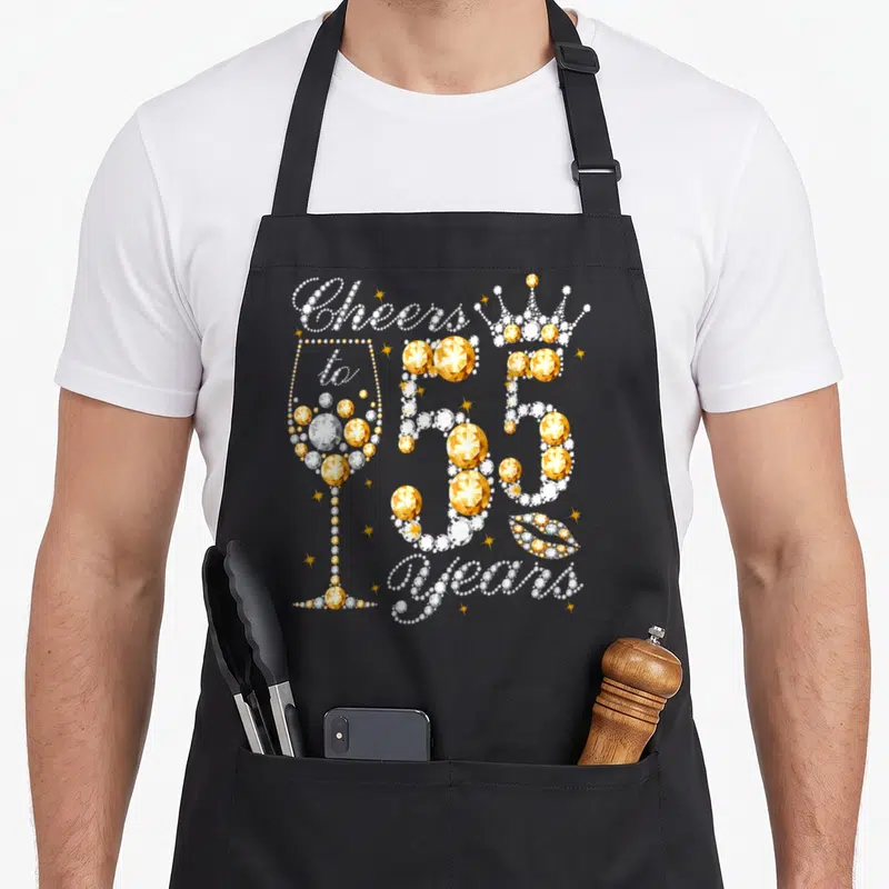 Cheer wine apron - Typography design with crown, lips, and glass spelling Cheers to 55 years in white and gold — shown on a black cotton canvas apron against a white background