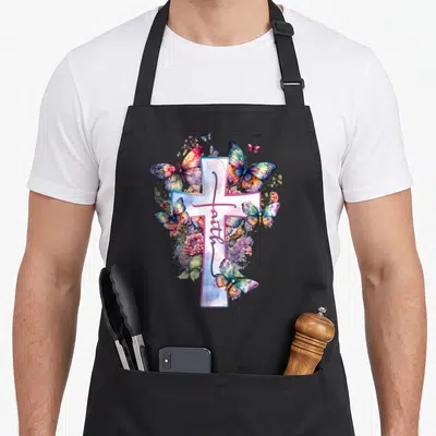 Floral cross apron - White cross with vertical faith text, watercolor butterflies and flowers in blue purple pink — shown on a black cotton canvas apron against a white background