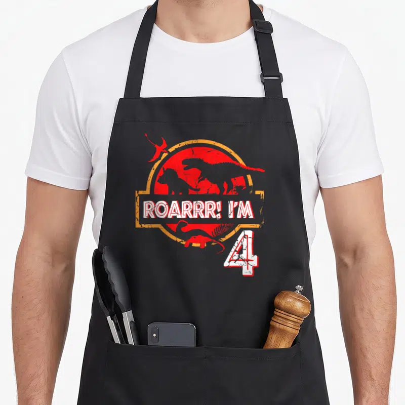 Birthday roar dinosaur apron - Red sun with dinosaur silhouettes and ROARRRR! I'M 4 text — shown on a black cotton canvas apron against a white background
