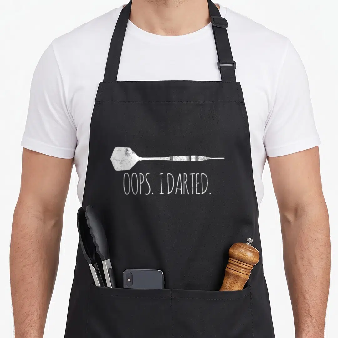 Funny darts apron - White distressed horizontal dart illustration above text OOPS. I DARTED — shown on a black cotton canvas apron against a white background