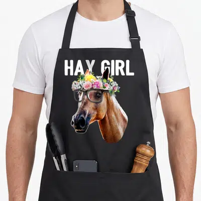 Hay floral apron - HAY GIRL text above horse head with flower crown and black glasses in brown white pink yellow green — shown on a black cotton canvas apron against a white background