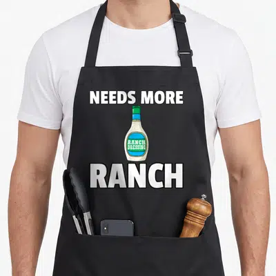 Condiment ranch dressing apron - Cartoon ranch dressing bottle with needs more text in white green and blue — shown on a black cotton canvas apron against a white background