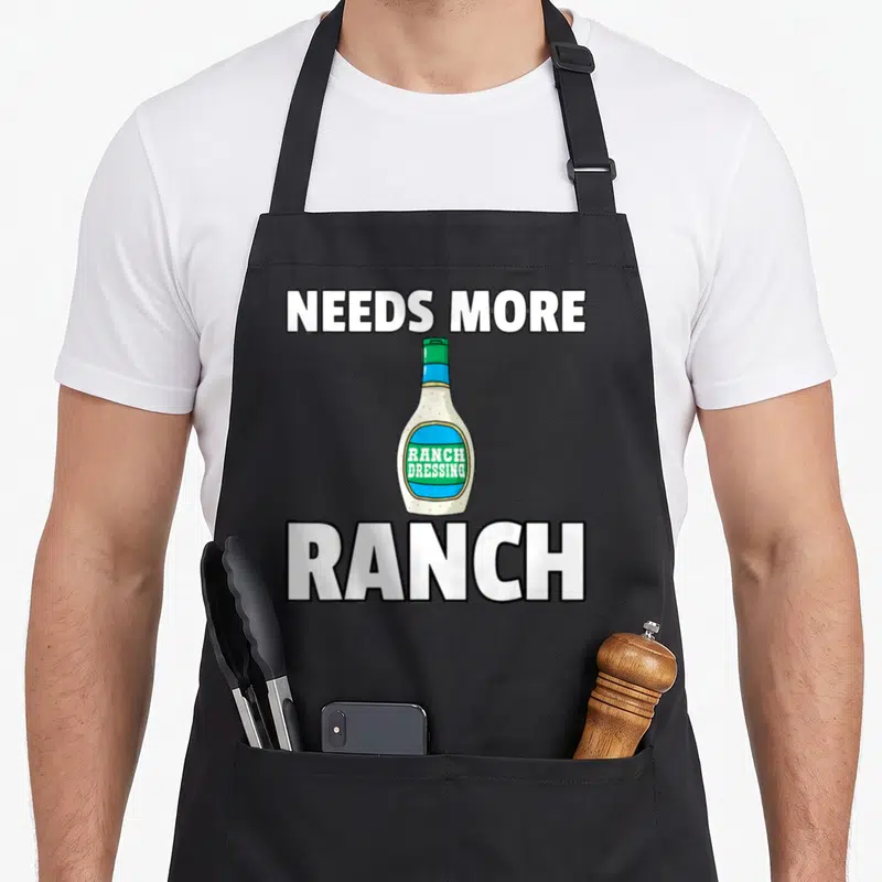 Condiment ranch dressing apron - Cartoon ranch dressing bottle with needs more text in white green and blue — shown on a black cotton canvas apron against a white background