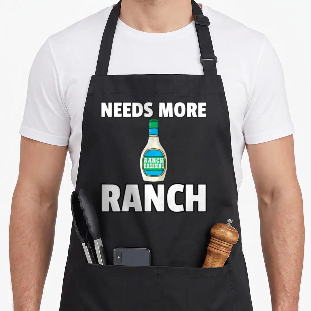 Condiment ranch dressing apron - Cartoon ranch dressing bottle with needs more text in white green and blue — shown on a black cotton canvas apron against a white background