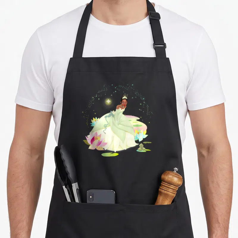 Princess frog aprons - Princess Tiana in green gown with pink yellow blue lotus flowers and small frog — shown on a black cotton canvas apron against a white background
