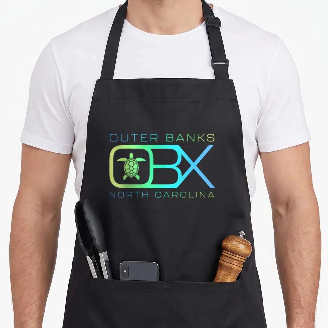 Vacation outer banks apron - Gradient OBX logo containing a sea turtle with blue and green text reading Outer Banks — shown on a black cotton canvas apron against a white background