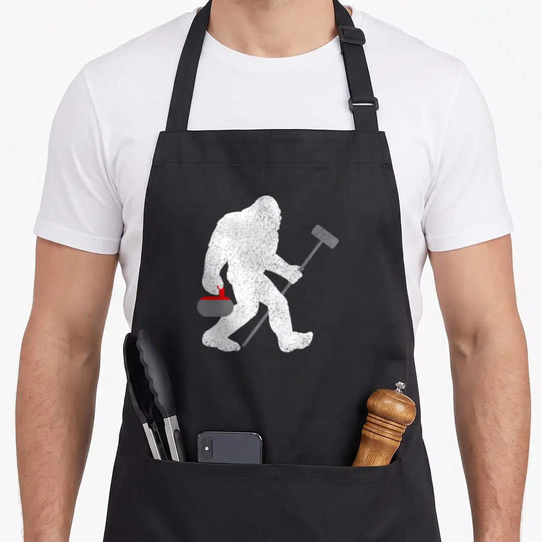 Curling sasquatch bigfoot apron - White distressed bigfoot silhouette walking right holding red curling stone and grey — shown on a black cotton canvas apron against a white background