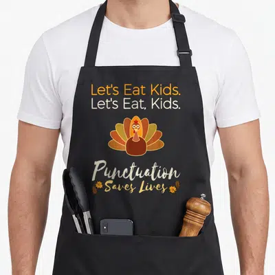 Grateful let's eat apron - Cartoon turkey with Let's Eat Kids text and two autumn leaves in orange and brown — shown on a black cotton canvas apron against a white background