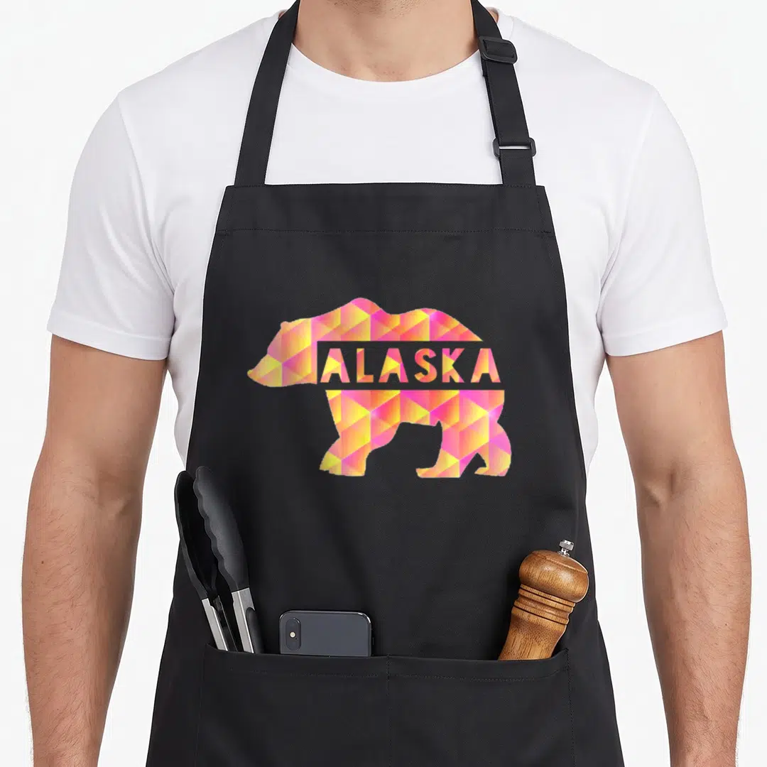 Proud bear apron - Geometric bear silhouette in pink yellow orange white with ALASKA text across chest — shown on a black cotton canvas apron against a white background