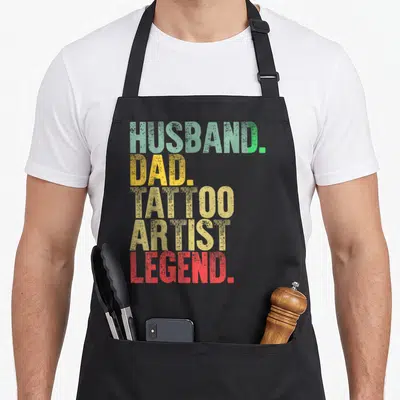 Husband tattoo aprons - Stacked text HUSBAND DAD TATTOO ARTIST LEGEND in green yellow beige red vintage font — shown on a black cotton canvas apron against a white background