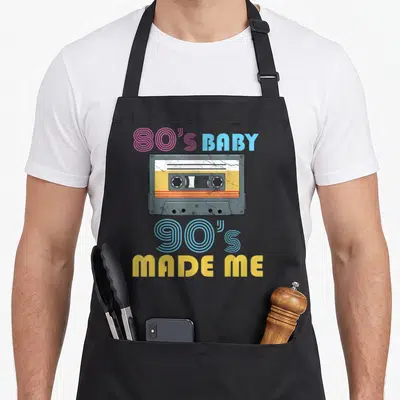 Outfit hip apron - Grey cassette tape with pink and blue 80s BABY 90's MADE ME text and yellow accents — shown on a black cotton canvas apron against a white background