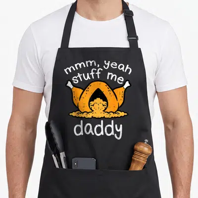 Funny turkey thanksgiving apron - Cartoon roasted turkey with mmm yeah stuff me daddy text in orange white and yellow — shown on a black cotton canvas apron against a white background