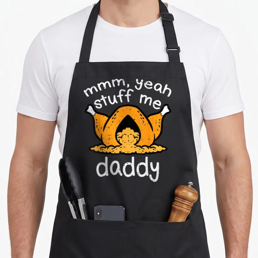 Funny turkey thanksgiving apron - Cartoon roasted turkey with mmm yeah stuff me daddy text in orange white and yellow — shown on a black cotton canvas apron against a white background