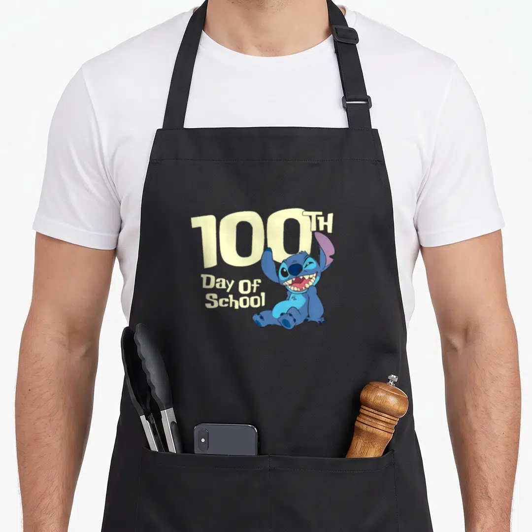 100th stitch apron - Blue cartoon character sitting next to 100TH Day Of School text in pale yellow — shown on a black cotton canvas apron against a white background