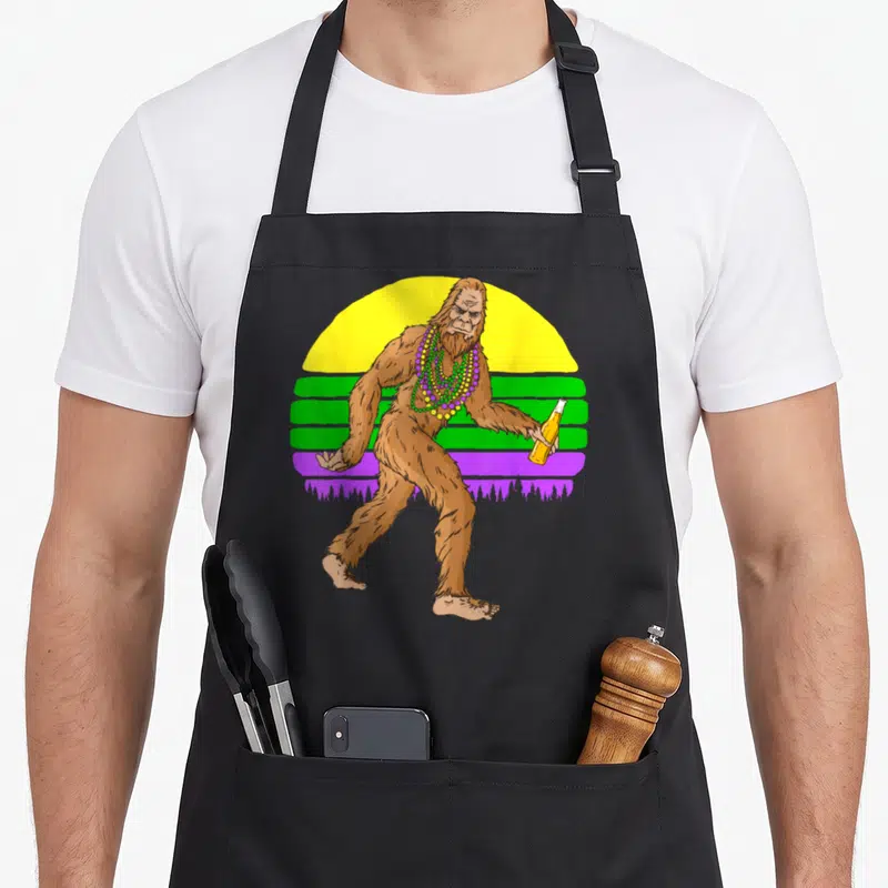 Bigfoot fat chef apron - Brown cartoon Bigfoot holding a bottle wearing purple green and yellow beads against a striped — shown on a black cotton canvas apron against a white background