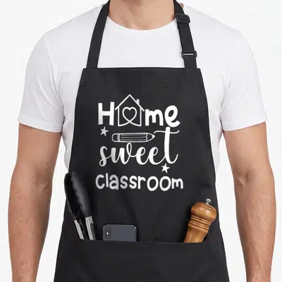 Sweet school classroom aprons - White stacked text Home Sweet Classroom with pencil and heart house icon — shown on a black cotton canvas apron against a white background