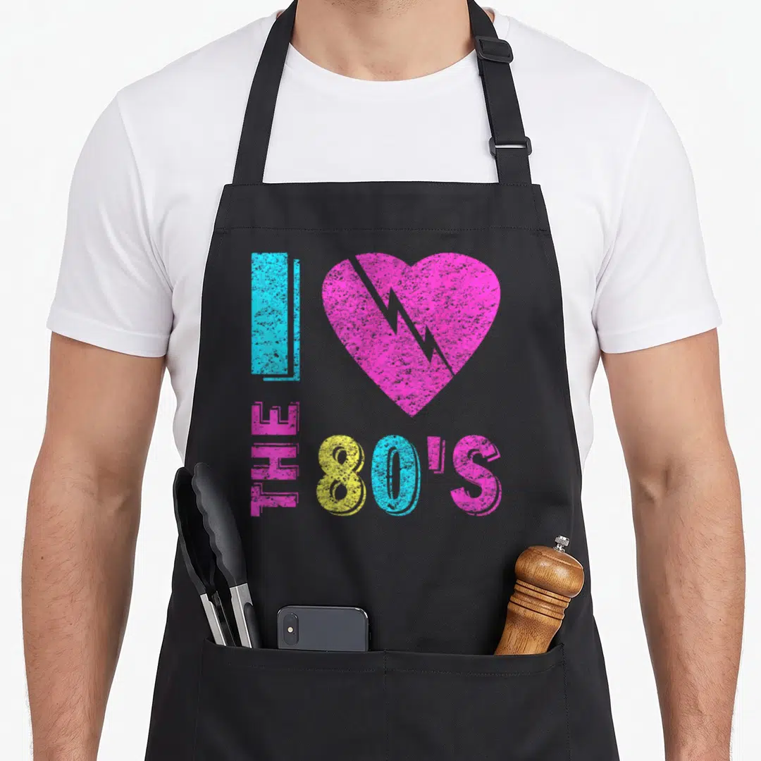 90s costume 80s apron - Distressed I love The 80s text with cracked pink heart and blue yellow letters — shown on a black cotton canvas apron against a white background