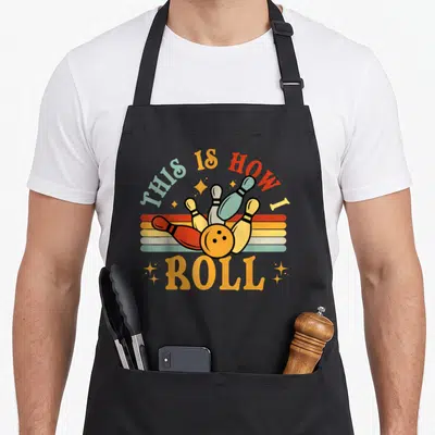 Funny bowling roll apron - Retro THIS IS HOW ROLL text with yellow red teal pins and horizontal stripes — shown on a black cotton canvas apron against a white background