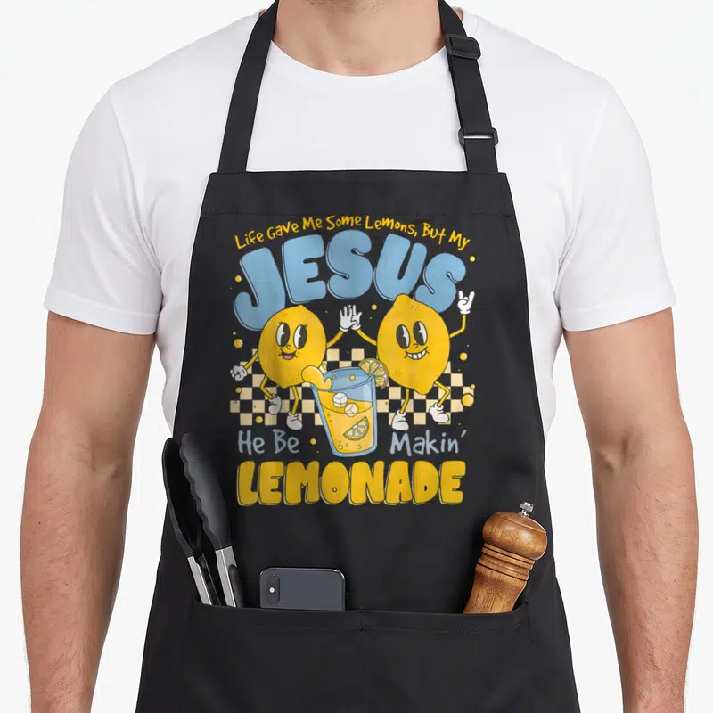 Gave lemonade apron - Two cartoon lemons high-fiving over lemonade glass with JESUS LEMONADE text — shown on a black cotton canvas apron against a white background