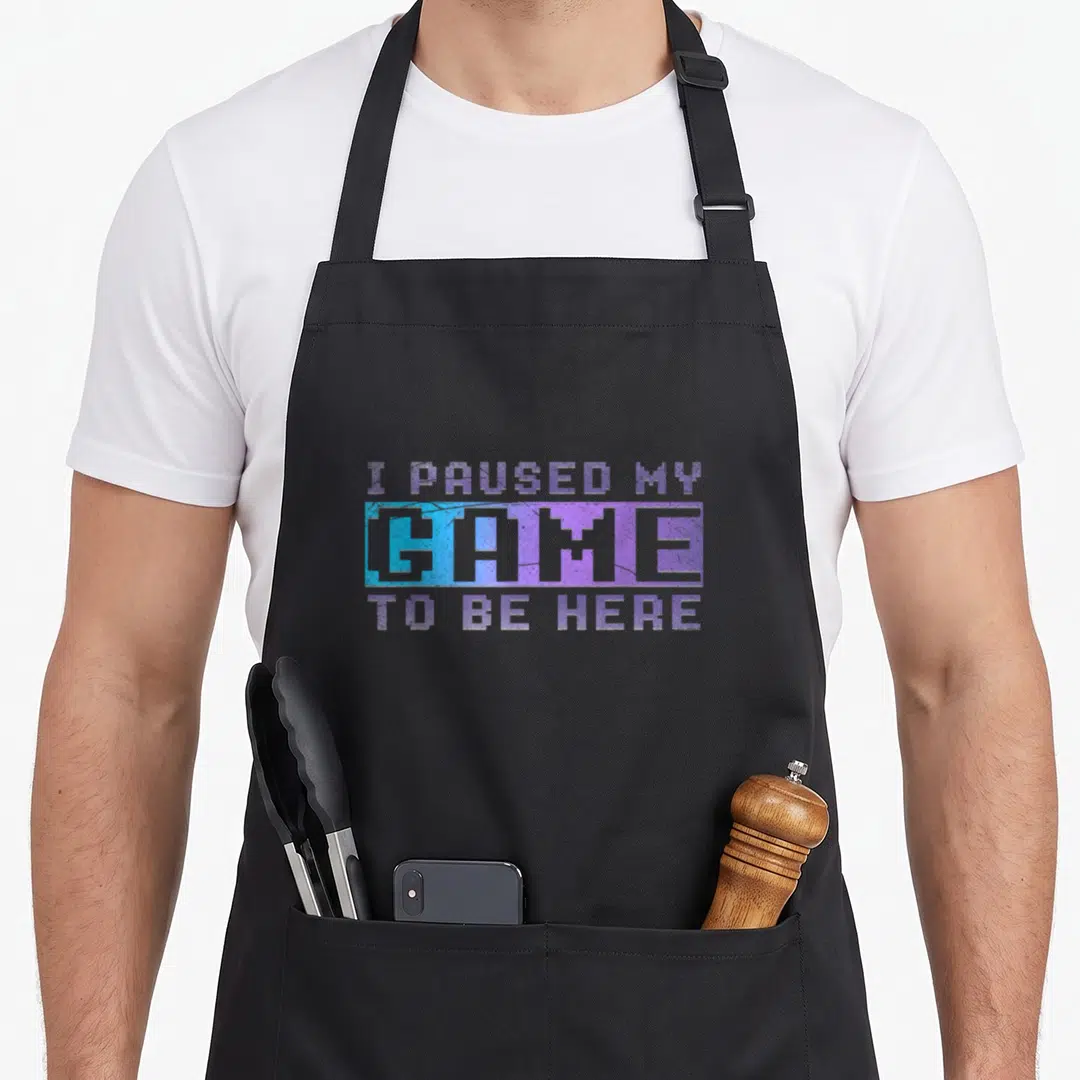Ich gaming apron - Purple and blue pixelated text reading I Paused My Game To Be Here stacked in three lines — shown on a black cotton canvas apron against a white background