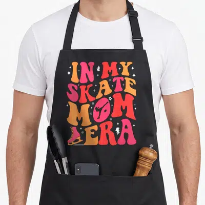 Skate mom era apron - Pink and orange text reading In My Skate Mom Era with figure skater silhouette in O and ice skate — shown on a black cotton canvas apron against a white background