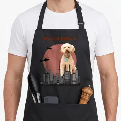 Doodlezilla goldendoodle apron - Giant cream dog behind black skyline and red sun with DOODLEZILLA text — shown on a black cotton canvas apron against a white background