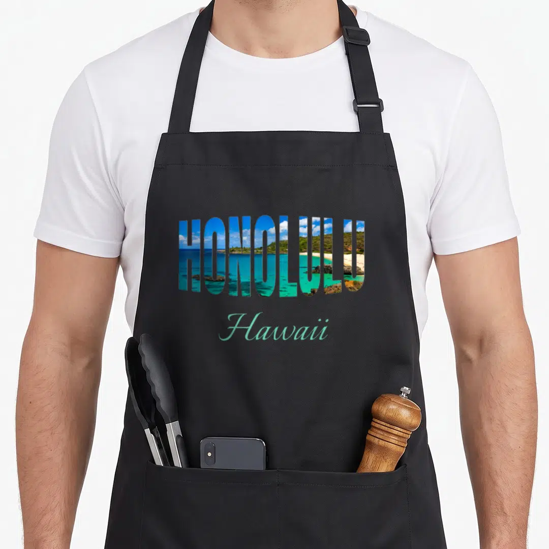 Honolulu sightseeing apron - HONOLULU text filled with beach landscape and turquoise Hawaii script — shown on a black cotton canvas apron against a white background