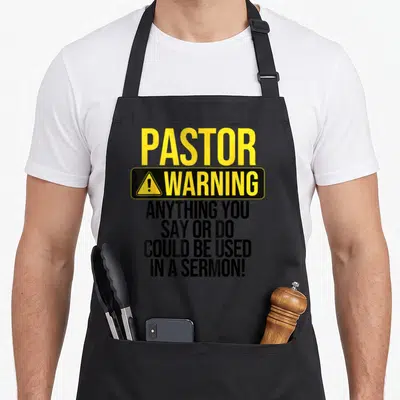 Pastor religious aprons - Yellow and black warning sign typography reading Pastor Warning Anything You Say Or Do Could — shown on a black cotton canvas apron against a white background