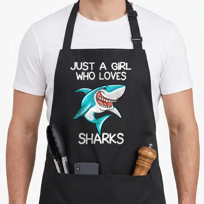 Cute shark apron - Blue cartoon shark with open mouth and white text Just A Girl Who Loves Sharks — shown on a black cotton canvas apron against a white background