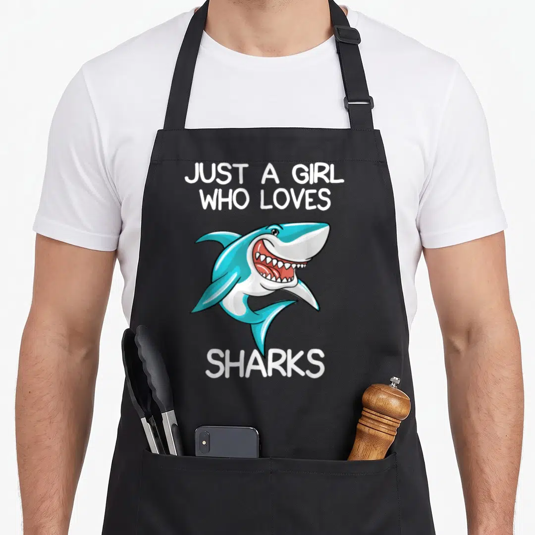 Cute shark apron - Blue cartoon shark with open mouth and white text Just A Girl Who Loves Sharks — shown on a black cotton canvas apron against a white background