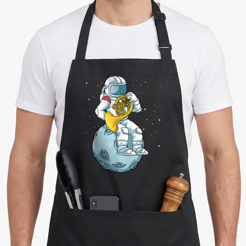 Brass french apron - Cartoon astronaut playing french horn on a blue cratered moon with scattered stars — shown on a black cotton canvas apron against a white background