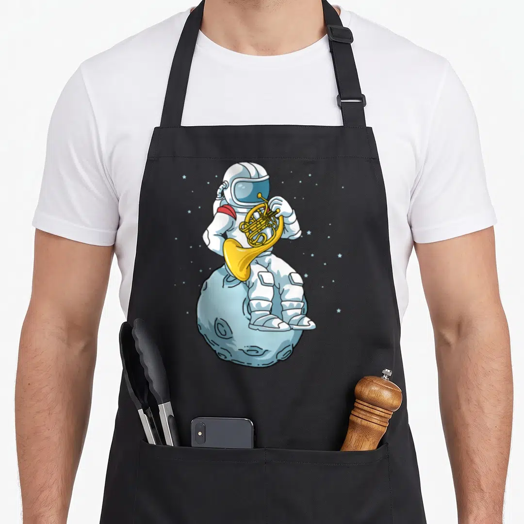 Brass french apron - Cartoon astronaut playing french horn on a blue cratered moon with scattered stars — shown on a black cotton canvas apron against a white background