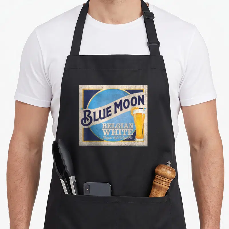 Coor moon retro apron - Blue Moon Belgian White logo with beer glass and orange slice on light blue circle — shown on a black cotton canvas apron against a white background