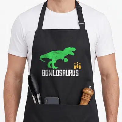 Bowler dinosaur apron - Green T-Rex holding bowling ball above BOWLOSAURUS text with three yellow pins — shown on a black cotton canvas apron against a white background