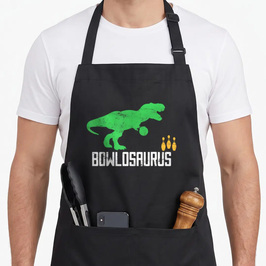 Bowler dinosaur apron - Green T-Rex holding bowling ball above BOWLOSAURUS text with three yellow pins — shown on a black cotton canvas apron against a white background