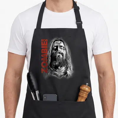 Zombie face apron - Grayscale zombie head photo with vertical red ROB ZOMBIE text on grey background — shown on a black cotton canvas apron against a white background