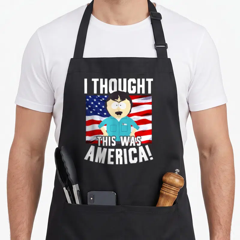 Randy cartoon apron - Cartoon man centered on waving flag with white text reading I thought this was america — shown on a black cotton canvas apron against a white background