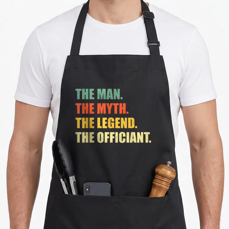 Officiant ordained minister wedding apron - Stacked text reading The Man The Myth The Legend The Officiant in green red — shown on a black cotton canvas apron against a white background