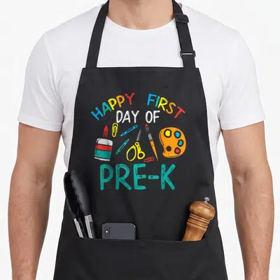 Pre prek kindergarten teacher apron - HAPPY FIRST DAY OF PRE-K text over cartoon scissors crayons and paint palette — shown on a black cotton canvas apron against a white background