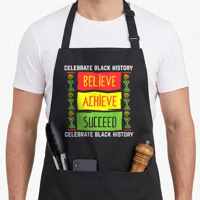 Believe political aprons - CELEBRATE BLACK HISTORY text with red yellow green BELIEVE ACHIEVE SUCCEED banners and sun — shown on a black cotton canvas apron against a white background