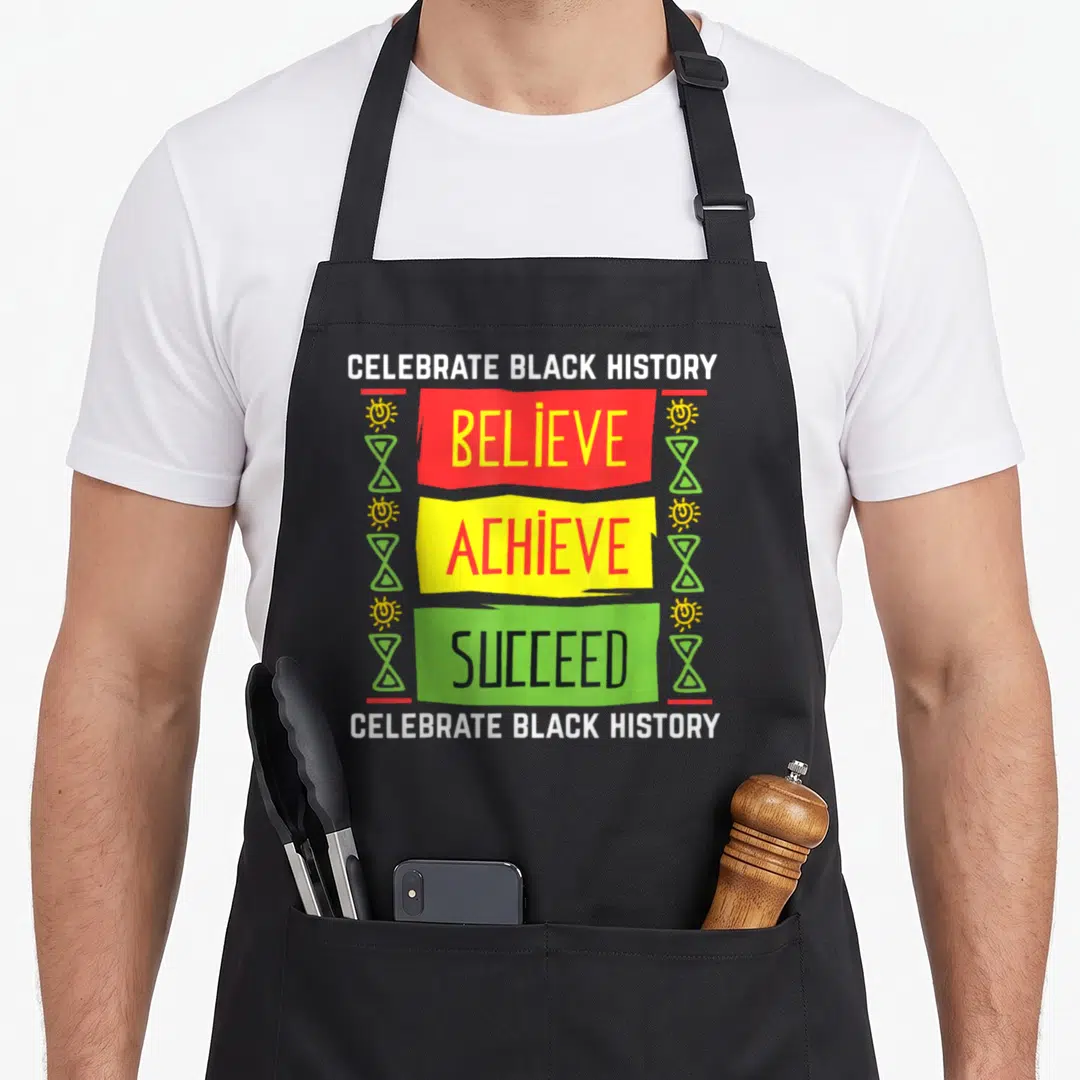 Believe political aprons - CELEBRATE BLACK HISTORY text with red yellow green BELIEVE ACHIEVE SUCCEED banners and sun — shown on a black cotton canvas apron against a white background