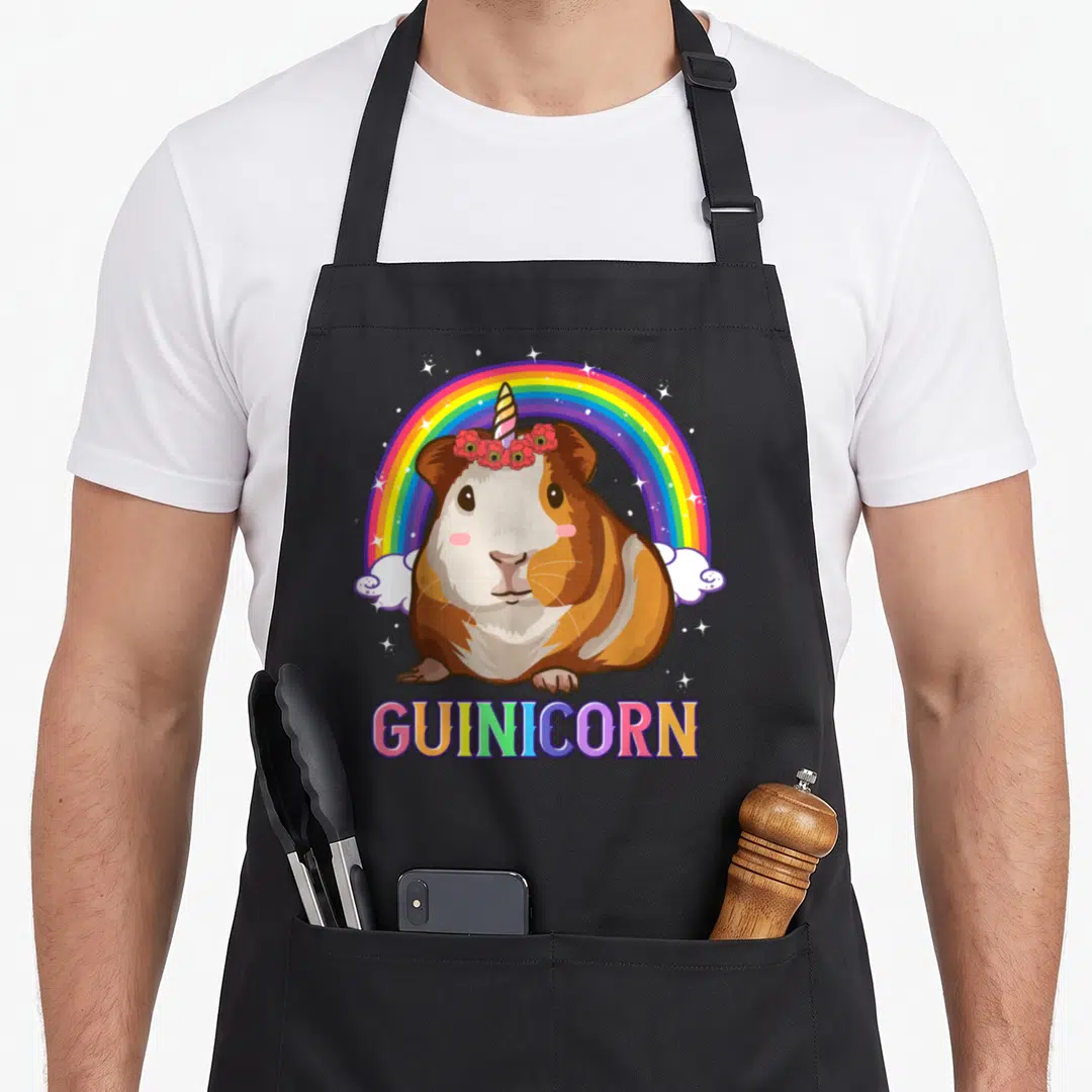 Guinea unicorn apron - Cartoon brown and white guinicorn with red flower crown under rainbow and GUINICORN text — shown on a black cotton canvas apron against a white background